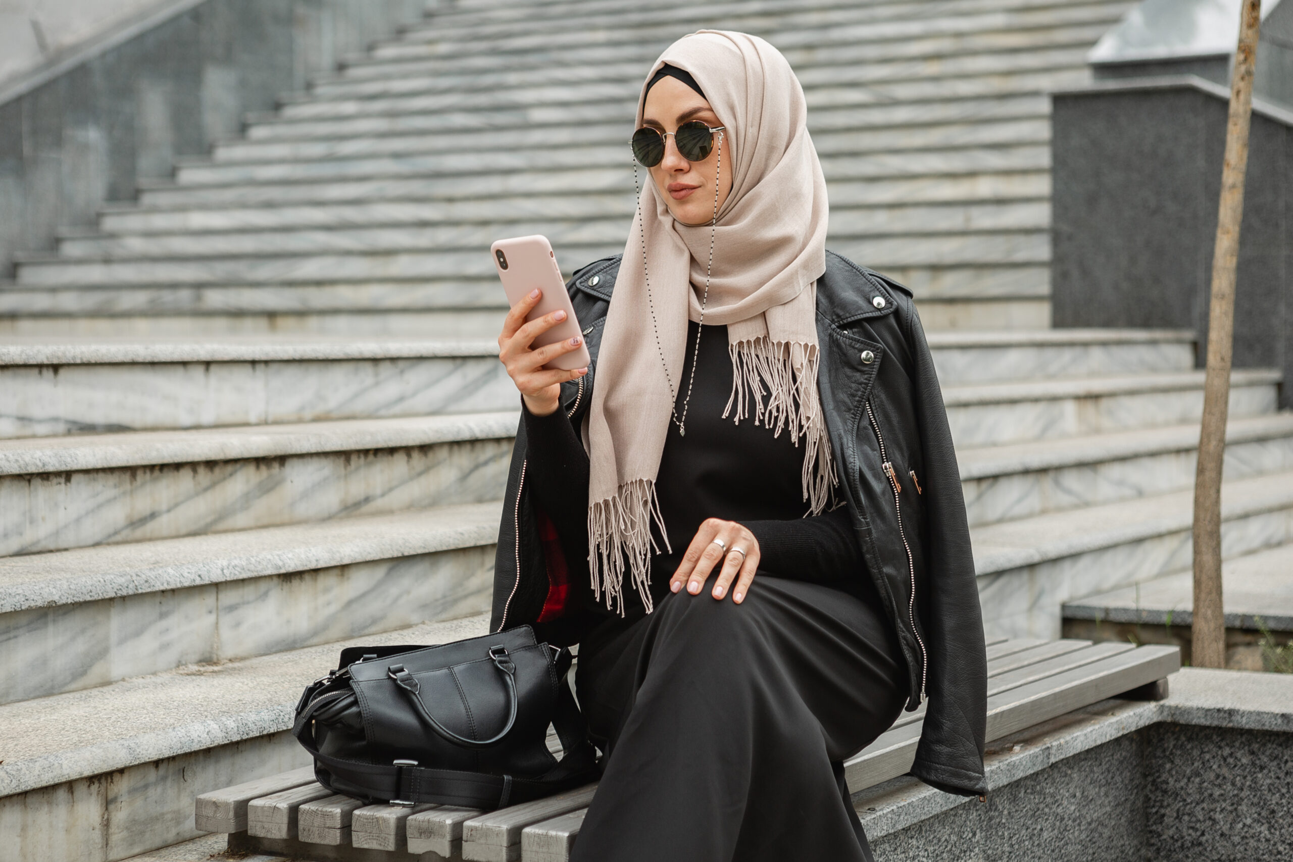 modern stylish muslim woman in hijab, leather jacket and black abaya walking in city street using smartphone, wearing sunglasses and bag, fashion style trend
